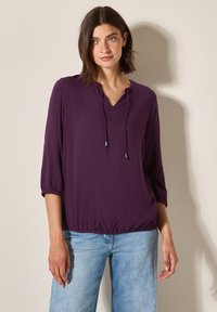 Blouse violette légère avec un col en V et un ourlet élastique, dotée de manches trois-quarts et d'un détail à cordon. À associer avec un jean bleu clair.