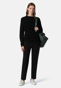 Schwarzer Strickpullover, schwarze Hose und weiße Sneaker mit roten Akzenten. Grünes Bag mit umschlagbarem Design und Logodetail. Glatte Textur.