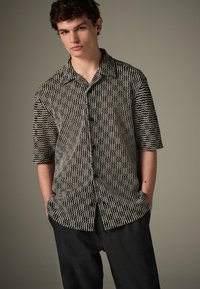 Camicia a maniche corte con motivo ondulato bianco e nero, con abbottonatura frontale, collo aperto e vestibilità rilassata. Paio di pantaloni neri.