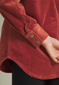 Detalle de camisa de pana roja que muestra prominentes costillas, puño con dos botones metálicos y dobladillo curvado con un ajuste ligeramente holgado.