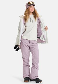 Strój narciarski składający się z białej i lawendowej kurtki, jasnego topu, fioletowych spodni, czarnych butów i przyciemnianych gogli narciarskich.