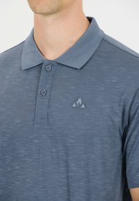 Blå pikétröja är gjord av ett texturerat tyg. Har en krage, två knappar och en liten triangel-logotyp på vänster sida. Korta ärmar.