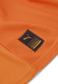 Maglietta da calcio arancione con tessuto testurizzato. Presenta un'etichetta blu scuro con scritte dorate che recitano "CALCIO AUTENTICO LICENZIATO" e un logo.