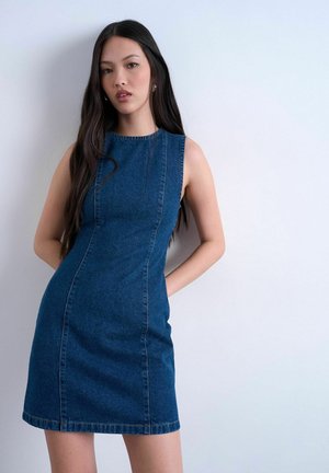 Jonge vrouw met lang donker haar, gekleed in een mouwloze blauwe spijkerjurk, staand tegen een eenvoudige lichtgrijze muur.