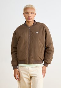Casaco bombardeiro castanho com fecho de zip, punhos e bainha canelados, e um pequeno logótipo branco, combinado com calças de tom claro e uma blusa verde menta.