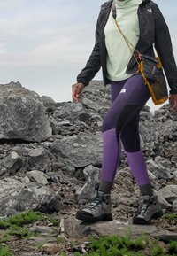 Chaqueta negra, top verde claro, leggings morados con acentos negros, botas de senderismo grises. Modelo caminando sobre terreno rocoso.