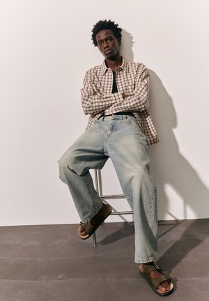 Jonge man met een geruit overhemd, lichtblauwe wijde spijkerbroek en bruine sandalen zit op een metalen kruk tegen een witte muur.