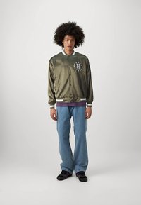 Olive rohelise satiinist pommitaja jakk valge äärise, rinnal oleva logoga ja tähtedega. Ühildatud siniste teksade ja mustade tossudega, millel on lilla detailid.