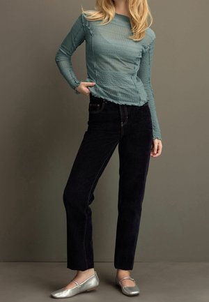 Femme portant un haut à manches longues texturé de couleur bleu sarcelle, un pantalon noir et des ballerines argentées, debout devant un fond marron uni.