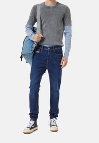 Grå kortärmad T-shirt med blå långärmad undertröja, mörkblå jeans och vita sneakers med mönstrade snörningar. Håller en blå ryggsäck.