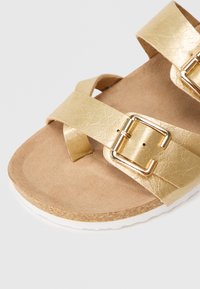 Χρυσά σανδάλια από faux δέρμα με δύο διακριτές λωρίδες, που διαθέτουν μεταλλικές αγκράφες, μαλακή μπεζ σόλα και υφασμάτινη φελλού με λευκή ακμή.