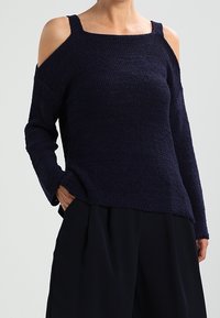 Mörkblå stickad tröja med off-shoulder-design, långa ärmar och en texturerad yta. Bärs med lösa svarta byxor som har en avslappnad passform.