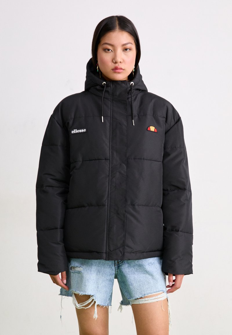Doudoune noire avec capuche, arborant un logo sur la poitrine gauche, des finitions en métal argenté et des poches latérales. Portée sur un short en denim déchiré.
