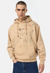 Beige kapucnis pulcsi elülső zsebbel, zöld és narancs fröccsenés mintákkal, valamint fekete alapon egyedi grafika a mellkasán.