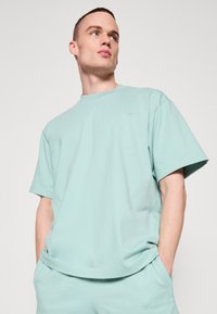 Tricou din bumbac de culoare verde mentă deschisă, cu guler rotund, mâneci scurte și un logo mic, embossat pe piept. Textură moale, croială lejeră.