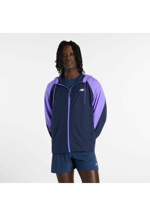 Mand med dreadlocks iført en navy- og lilla lynlåsjakke og navy shorts, stående med hænderne bag ryggen mod en hvid baggrund.