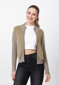 Θολό μπεζ και γκρι ζακέτα με φερμουάρ, με ριμπ μανίκια, κοντή γραμμή και αντίθετες υφές. Συνδυασμένη με μαύρο ψηλόμεσο παντελόνι.