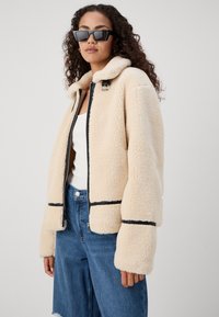 Beige faux shearling jacket with a zip front, black trim details, and a collar. Worn over a white tank and blue denim jeans.