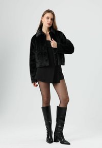 Veste courte en fausse fourrure noire avec col, fermeture à boutons et manches longues. Portée sur un haut noir et une jupe, assortie à des bottes noires montantes.