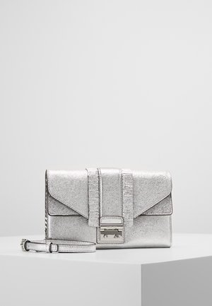 Pochette métallique argentée avec une boucle frontale, des bandes verticales décoratives et une bandoulière amovible sur une surface d’exposition blanche.