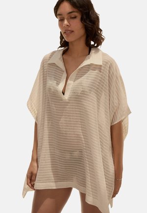 Femme porte un paréo ample, transparent beige à rayures avec des manches courtes et un col blanc par-dessus un maillot de bain noir, debout devant un fond uni.