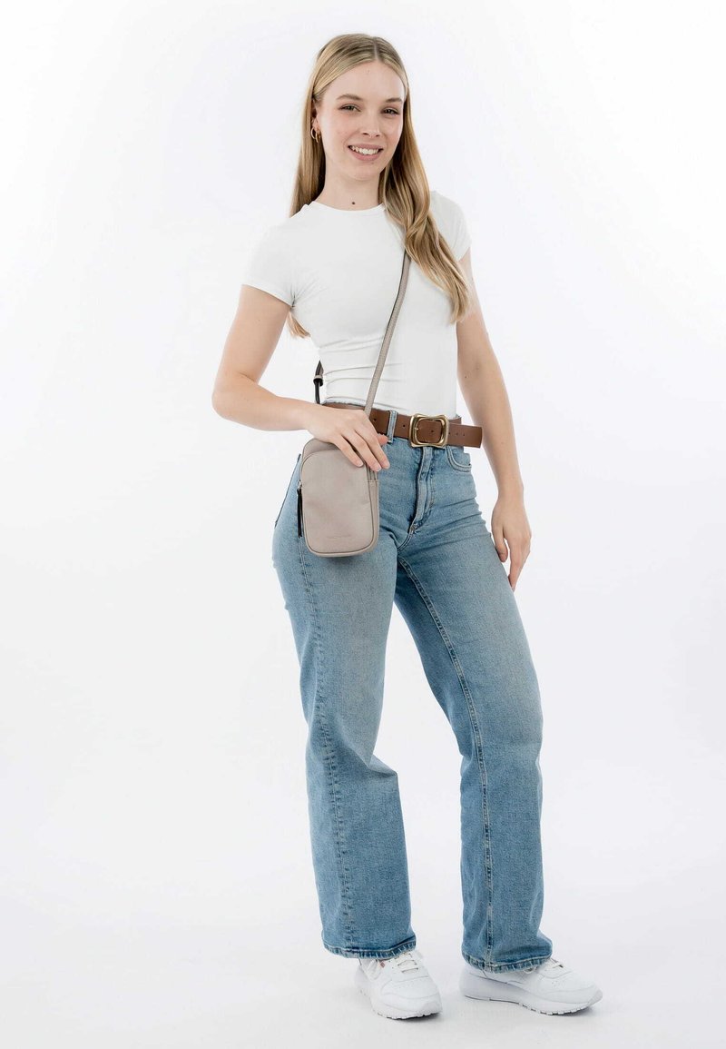 Ljust grå axelväska med en slät textur, bärs över en vit skjorta och blå jeans. Modellen står med ett brunt bälte och vita sneakers.