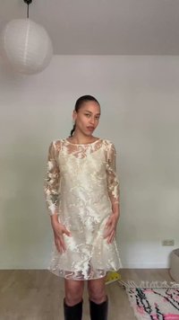 Femme portant une robe beige transparente à motifs avec des bottes noires montantes, debout à l'intérieur contre un mur uni.