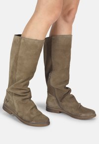 Bottes en daim brun jusqu'aux genoux avec des semelles plates et des fermetures éclair sur le côté, portées sur des jambes nues, montrant un léger froissement sur le matériau.