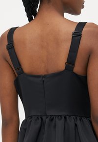 Melna kleita ar pieguļošu augumu un plisētu svārku. Iespējamas regulējamas siksnas un slēptais rāvējslēdzis aizmugurē. Gluda auduma tekstūra.