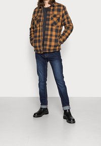 Brun och svart rutigt flanelljacka med två bröstfickor, bärs över en mörk skjorta, i kombination med mörka jeans och svarta ankelstövlar.