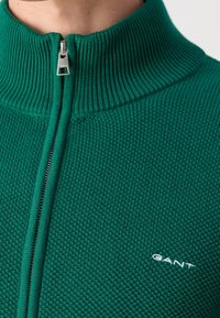 Gros plan d'un pull zippé vert texturé avec un col côtelé et un petit logo blanc GANT sur la poitrine.