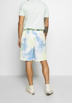 Mężczyzna w jasnoniebieskich i żółtych krótkich spodenkach tie-dye, białej koszulce, białych wysokich trampkach, stojący tyłem do aparatu na białym tle.