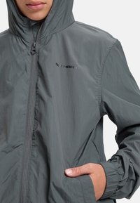 Grå lättviktsjacka med huva, dragkedja fram, elastiska ärmslut och en logotyp på bröstet. Strukturerad tyg med en slät yta.