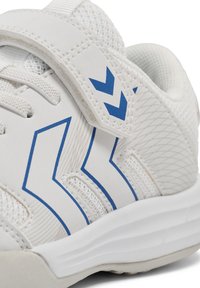 Bottine de sport blanche avec des motifs en chevron bleu, panneaux en maille, et une sangle Velcro sur des lacets blancs, présentée en gros plan de profil.