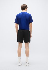 Modré tričko s krátkym rukávom s textúrovaným vzorom, v kombinácii s čiernymi šortkami. Model má na sebe biele športové ponožky a tenisky.