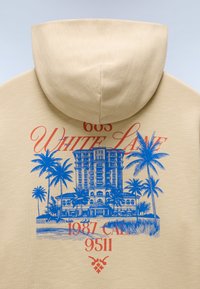 Sweat à capuche beige avec une illustration bleue d'un bâtiment entouré de palmiers et un texte rouge indiquant "605 White Lane 1987 CAL 9511".