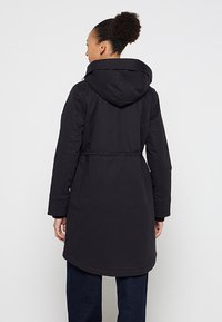 Zwarte capuchonjack met een getailleerde taille, lange mouwen en een licht gebogen zoom. Gemaakt van glad, waterafstotend materiaal.