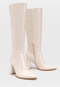 Bottines en cuir beige montantes jusqu'au genou, avec un bout pointu et un talon carré. Elles possèdent une fermeture éclair sur le côté et une texture lisse.