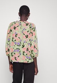 Blouse fleurie en tissu vert clair. Présente de grandes roses roses et des fleurs violettes, avec des manches bouffantes et un détail de col à volants.