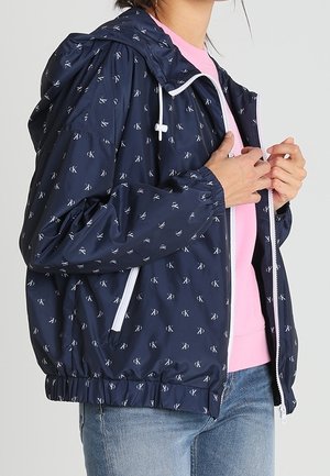 Persoon draagt een donkerblauwe capuchonjas met witte rits en klein wit logopatroon, roze shirt en blauwe spijkerbroek, en stelt de jasrits af.