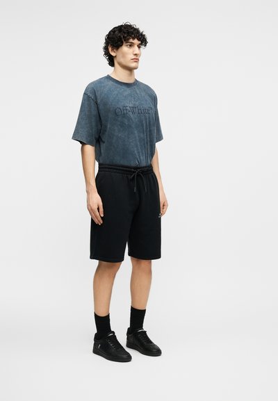 Homme aux cheveux bouclés portant un T-shirt Off-White bleu délavé, un short noir à cordon, des chaussettes noires et des baskets noires, debout devant un fond uni.