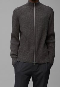 Ciemnoszary sweter prążkowany z wysokim kołnierzem, pełnym zamkiem błyskawicznym i teksturowanymi detalami na szwach ramion, noszony na ciemnych spodniach.