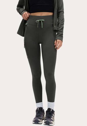 Person iført mørkegrønne leggings med sidelomme, sort crop top, matchende jakke, hvide sokker og sorte træningssko, stående.