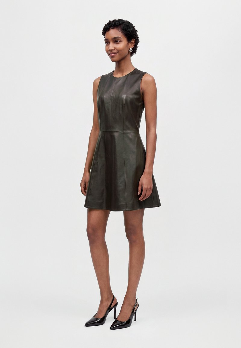 Une femme porte une mini-robe en cuir foncé sans manches et des escarpins noirs pointus sur un fond clair et uni.