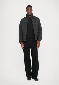 Man wearing black quilted jacket over black sweater, black wide-leg pants, and black shoes, standing against a plain white background.