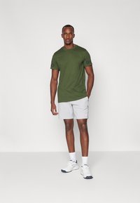 Olivengrønn crew-neck t-skjorte med svart logo på ermene, paret med lysegrå shorts og hvite joggesko. Flat tekstur, avslappet passform.