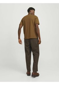Brun kortærmet t-shirt, parret med mørkebrune bukser. Skjorten har en løs pasform; stoffet ser blødt og glat ud. Fodtøj: brune sko.
