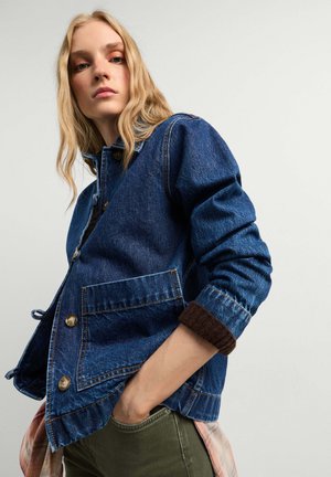 Junge Frau mit langen blonden Haaren, die eine blaue Jeansjacke und grüne Hosen trägt, steht vor einem schlichten hellen Hintergrund.