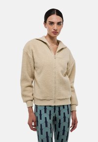 Veste beige zippée en tissu doux et texturé, avec col et manches côtelés. Présente une coupe décontractée et un design sans coutures.