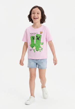 Barn med kort krøllet hår, der smiler, iført en lyserød Minecraft Creeper T-shirt, lyse denimshorts og hvide sneakers, går fremad.
